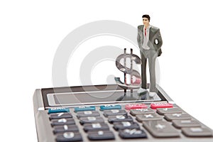 A businessman doll in a business suit stands on the glass of the calculator next to the dollar sign.
