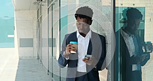 Businessman drinking coffee and using mobile phone leaning on wall of modern office building