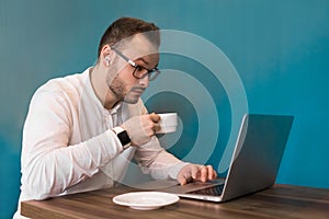 A businessman from Europe with glasses, with a beard and a smart watch on his hand drinks coffee and works in a laptop sitting at