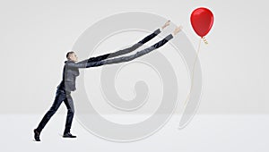 A businessman with extremely long arms trying to catch a red balloon with a gift gold ribbon.