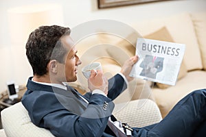 Businessman in formal suit sitting in armchair reading newspaper and drinking cup