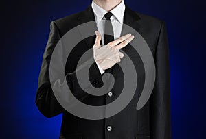 Businessman and gesture topic: a man in a black suit and white shirt, put his hand on his chest as a sign of a pistol on a dark bl