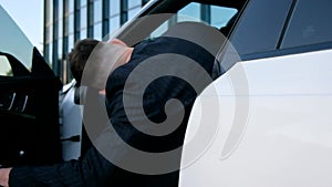 A businessman gets behind the wheel of a white car. The man opens the door and gets inside the car.