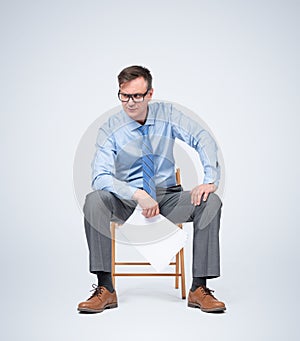 Businessman in glasses, a shirt and tie sits on a chair holding paper in his hands, on light blue background.