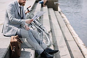 businessman in grey suit reading newspaper on stairs on quay with bicycle