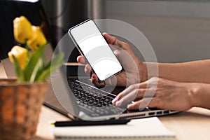 The businessman hand holding blank screen mobile phone during working on laptop computer at the home office.