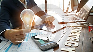 Businessman hand holding lightbulb with business documents and strategy word on office table and banking data on smart phone
