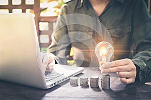 Businessman hand holding lightbulb on stack coins, and working with computer on workplace. Creative ideas concept of saving money