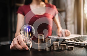 Businessman hand holding lightbulb with using laptop computer and money stack in office.