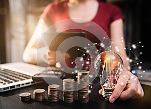 Businessman hand holding lightbulb with using laptop computer and money stack in office.