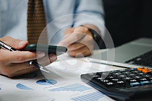 Businessman hand holding mobile phone and pen with graphs and charts business documents on desk. Calculator and computer for