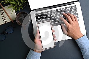 Businessman hand holding mobile phone and typing on laptop computer at office desk.