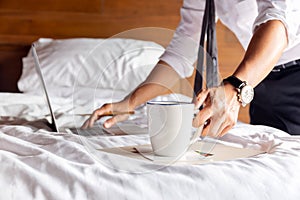 Businessman hand holding mug of coffee and working on laptop in bedroom.