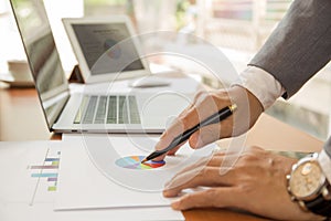 Businessman hand holding pen pointing to graph financial diagram with laptops and tablet on table.