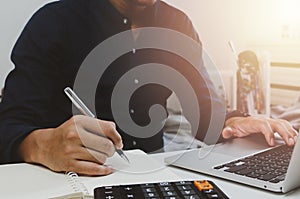 Businessman hand holding pen writing notebook and using keyboard computer laptop on desk