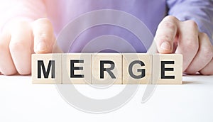 Businessman hand holding wooden cube block with MERGE business word on table background