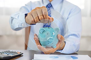 Businessman hand putting coin into blue piggy bank, saving money for future investment plan and retirement fund concept