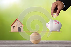 Businessman hand putting coin into piggy bank with model house on marble seesaw balancing on green background. Property investment