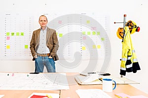 Businessman With Hands In Pockets Standing By Blueprint On Table