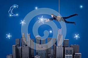 A businessman hanging on a rope above a night city and trying to catch a star in the sky.