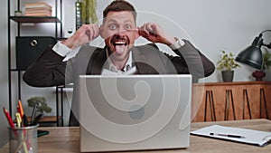 Businessman hiding behind laptop computer, making funny face, fooling around, putting his tongue out