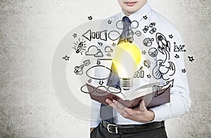 Businessman is holding a book with flying around business icons and a light bulb as a concept of the new business ideas.