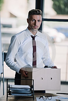 Businessman holding box while standing near workplace and looking at camera