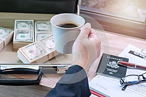 businessman holding cup of hot coffee with banknote on the bag and glasses , passport, pen and document on wooden table