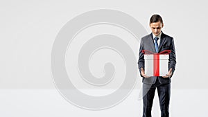 A businessman holding a gift box with a red bow in his arms looking down at it.