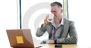 Businessman is holding glass of whiskey and looking at laptop due to stress of hard work due to poor economic conditions