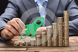 Businessman Holding Green Paper Rocket On Stacked Coins On Table