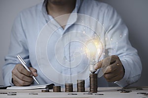 Businessman holding lightbulb which glowing on coin stacking and write down on notebook , Creative idea can make more money