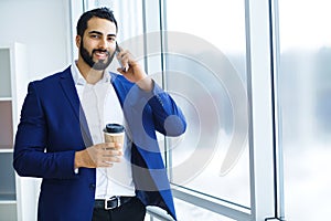 Businessman holding mobile phone and cup of coffee in office buildings in the background.