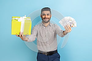 Businessman holding out wrapped present box and big fan of dollar banknotes, looking at camera.