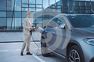 Businessman holding power supply cable at electric vehicle charging station.