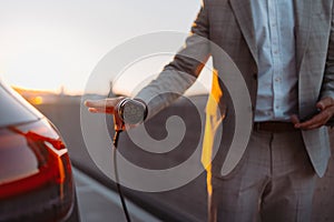 Businessman, holding power supply and charging his electric car during sunset. Concept of ecology tranport. photo