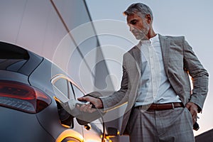 Businessman, holding power supply and charging his electric car during sunset. Concept of ecology tranport.