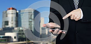 Businessman Holding Smartphone in Hand And Pointing Index Finger At The Phones Screen With Business City and Corporate Buildings