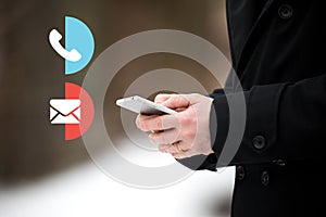 Businessman holding a smartphone mobile on a snowy winter day. Man in coat.With contact us call and email icons, customer service