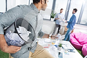 Businessman holding trash bucket with papers and looking at coworkers