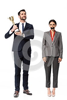 Businessman holding trophy near businesswoman with duct tape on mouth and medal isolated on white