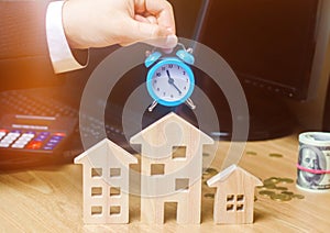 Businessman holds clock over wooden houses. Time to pay taxes. Mortgage. Payment of debts for real estate. Payment of utility