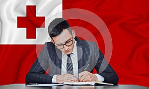 A businessman in a jacket and glasses sits at a table signs a contract against the background of a flag tonga, Nuku`alofa photo