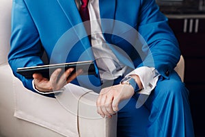 Businessman keep a digital tablet in hand whilst sitting on a sofa in a blue suit. on hand expensive mechanical watch