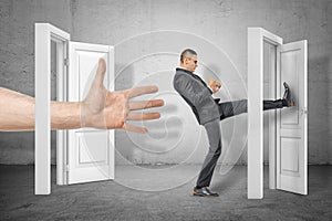 Businessman kicking a door and big hand appearing out of an open door on grey wall background