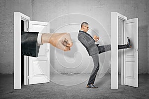 Businessman kicking door and big stretched fist appearing out of open door on grey wall background