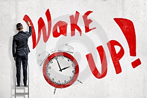 Businessman on a ladder facing the wall with red sign `Wake UP` and red alarm clock isolated on white background
