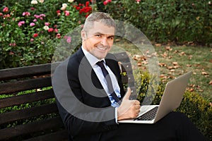 Businessman with laptop showing thumb-up gesture while sitting on bench outdoors
