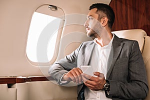 Businessman looking at airplane window while using smartphone in private jet