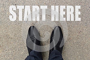 Businessman is looking down at his feet on a concrete floor with START HERE letters painted on the surface. Top view image of pers